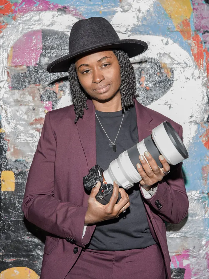 Portrait of Kash holding her camera and telephoto lens in front of colorful graffiti